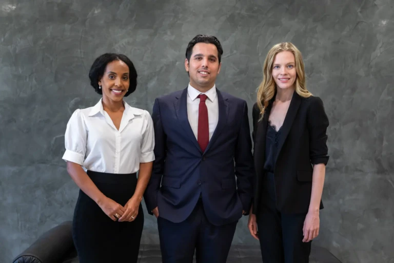 A Alex and two professionals standing against a gray textured wall. The two women and one man are in business attire, smiling confidently.
