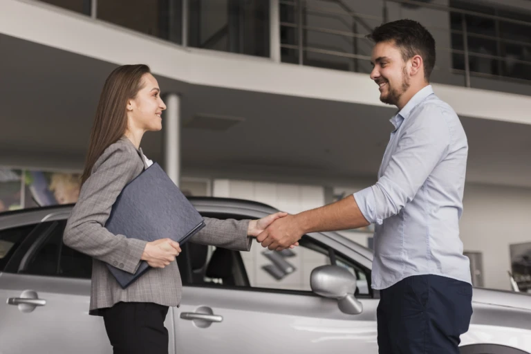 A woman in a gray suit shakes hands with a man in a blue shirt, smiling, standing in front of a silver car in a dealership, conveying professionalism.