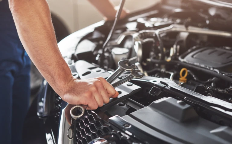 A mechanic's hand holding wrenches works on a car engine. Sunlight illuminates the open hood, emphasizing a focused and industrious atmosphere.