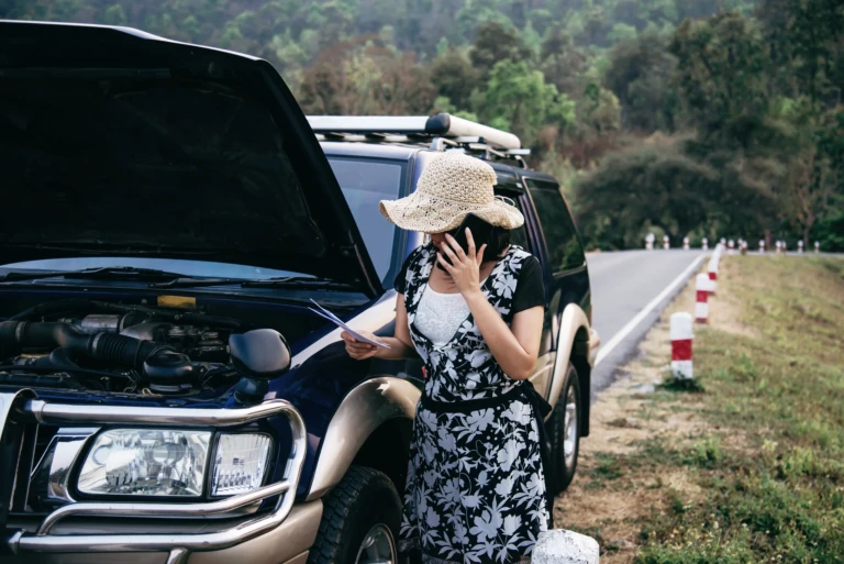A woman in a floral dress and straw hat stands beside a broken car with an open hood. She's on the phone, looking concerned, near a rural roadside.