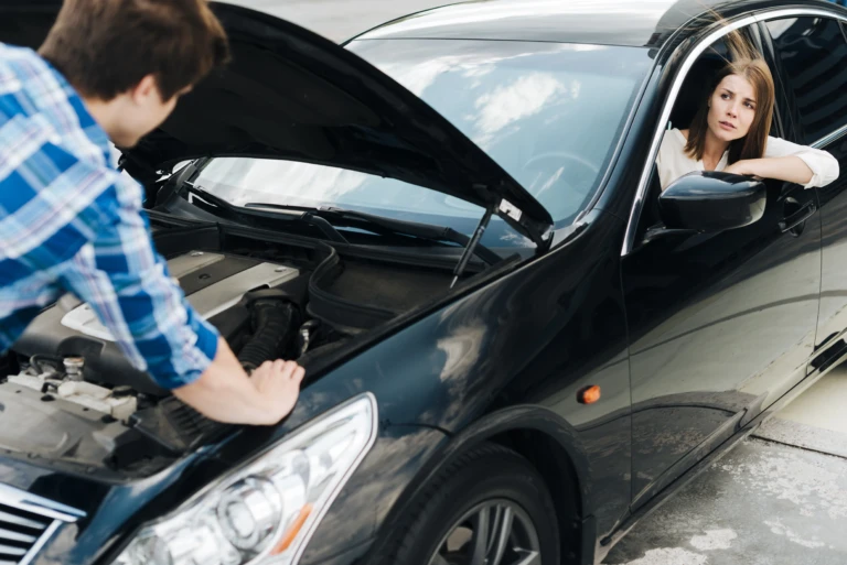 A woman looks concerned while sitting in a black car with the hood open. A man in a plaid shirt examines the engine, suggesting car trouble.