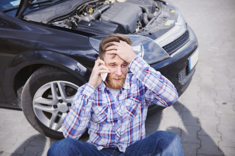 A man in a plaid shirt, sitting on the pavement, looks distressed while on the phone. A car with an open hood is in the background.