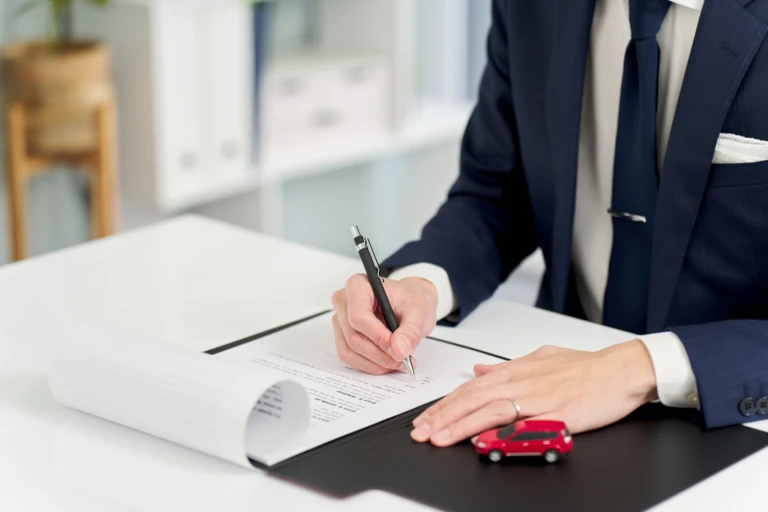 A person in a suit signs documents on a desk with a small red toy car. The scene suggests a professional setting, possibly related to car sales or loans.