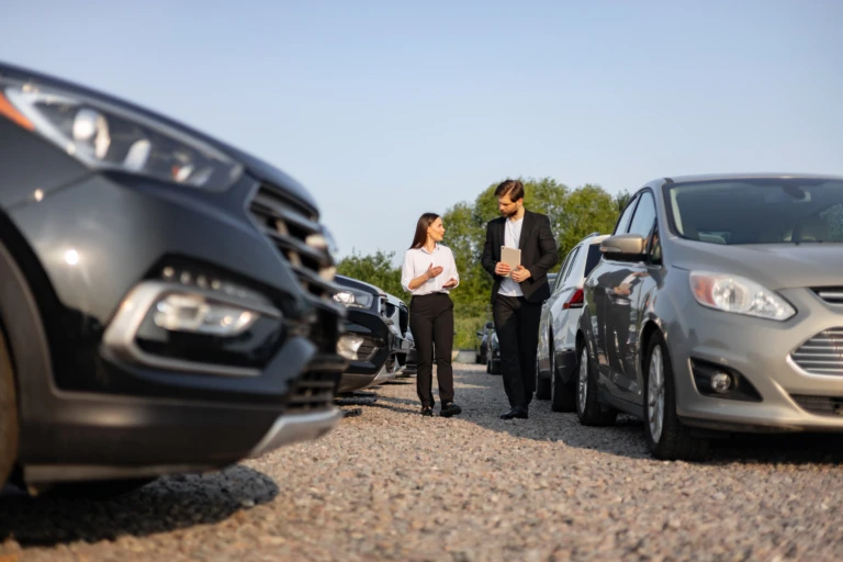 A man and a woman walk between rows of parked cars on a gravel lot. They are engaged in conversation, suggesting a sales or consultation setting.