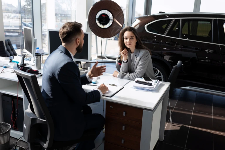 A man and woman are engaged in a discussion at a dealership desk. A car is visible through the window behind them. The atmosphere is professional.