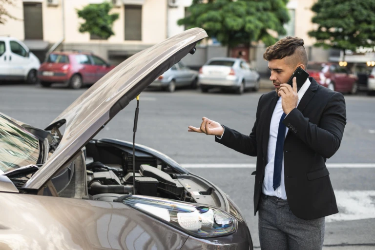 Man in a suit stands by a car with the hood open, talking on a phone and gesturing in frustration. Background shows parked cars and buildings.