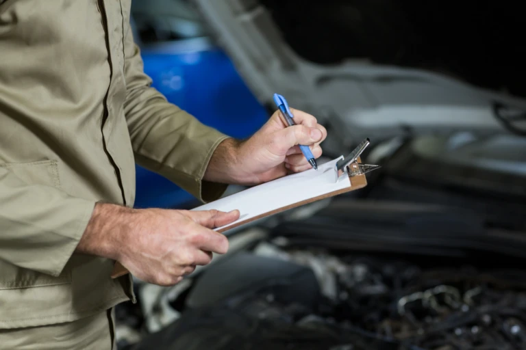 Mechanic in beige uniform writes on a clipboard in front of an open car hood, focusing on inspection. The atmosphere is professional and attentive.