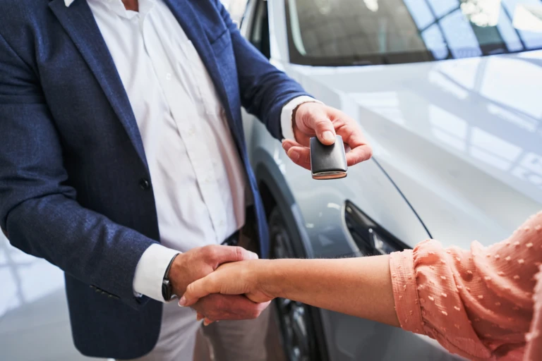 Two people shake hands while exchanging car keys in front of a sleek silver vehicle. The scene conveys a sense of agreement and new beginnings.